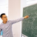 A male teacher points at equations on a blackboard during a lesson in a classroom setting.